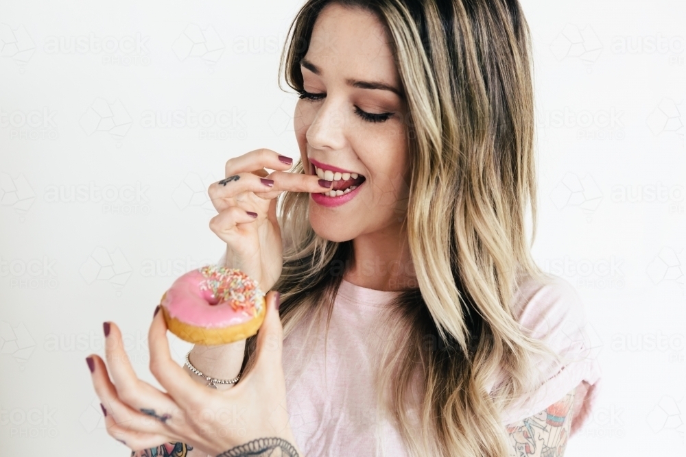 Girl licking her finger and about to eat a pink iced donut - Australian Stock Image