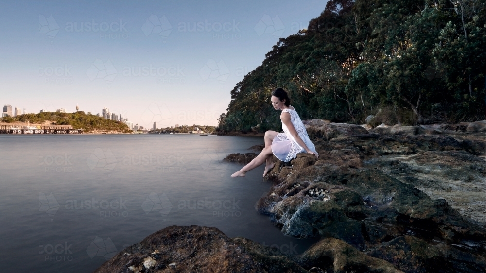 Girl peacefully relaxing by the water's edge in a white dress - Australian Stock Image