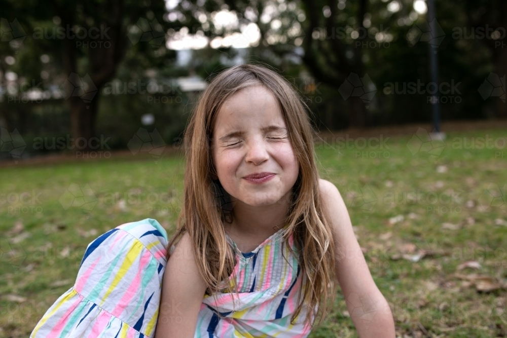 Girl pulling silly face with eyes closed sitting outside on grass - Australian Stock Image