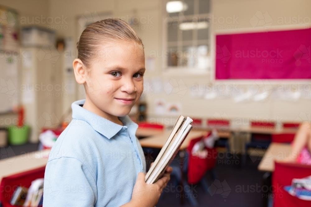 Girl school student in a classroom holding a set of books looking at camera - Australian Stock Image