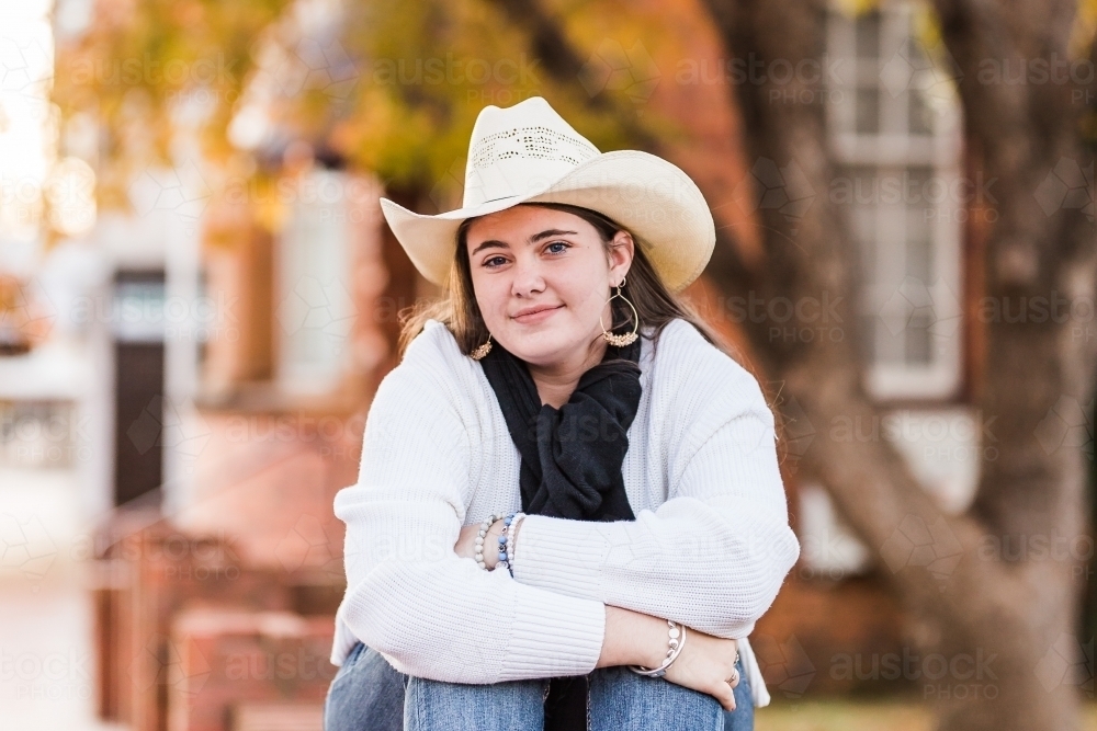 Girl sitting on brick wall hugging knees wearing hat smiling - Australian Stock Image