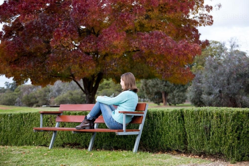 Girl sitting on park bench with autumn tree and hedge - Australian Stock Image