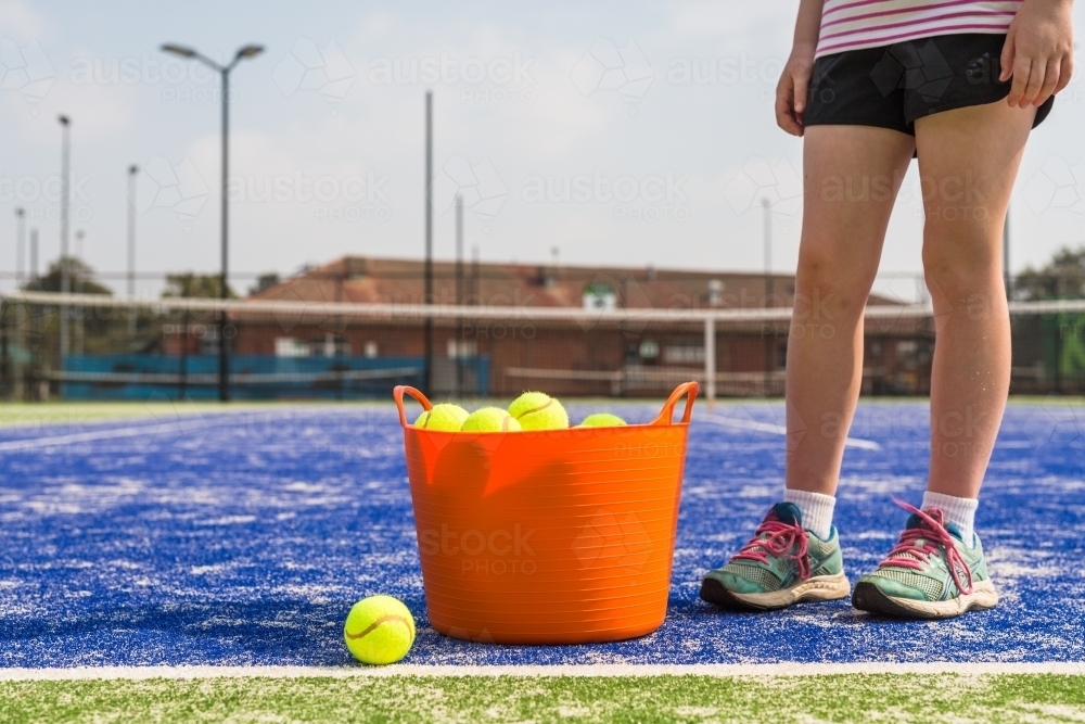 Girl standing next to an orange basket of tennis balls on a blue tennis court - Australian Stock Image