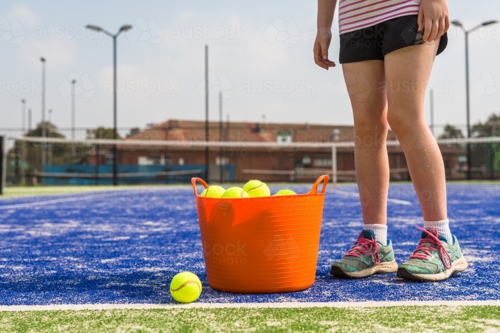 Girl standing next to an orange basket of tennis balls on a blue tennis court - Australian Stock Image