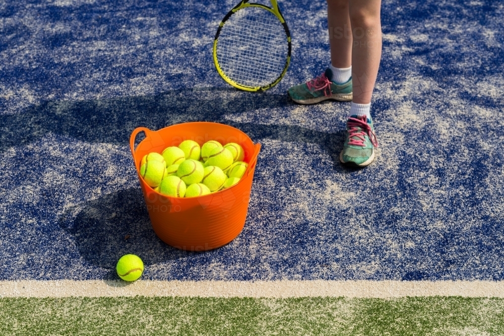 Girl standing next to an orange basket of tennis balls on a blue tennis court - Australian Stock Image