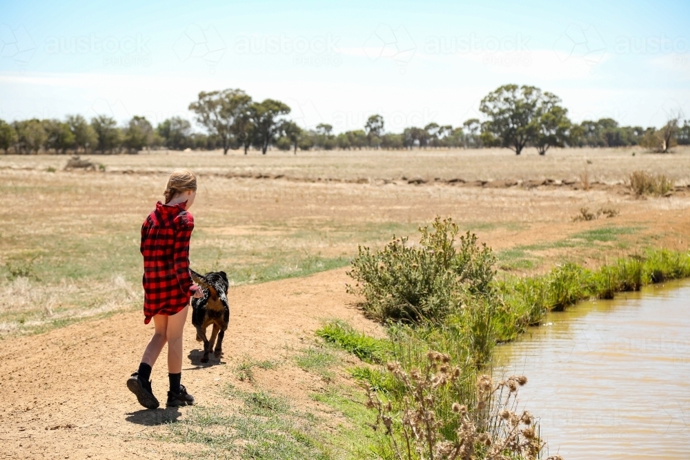 Girl walking alongside irrigation channel on farm with dog - Australian Stock Image