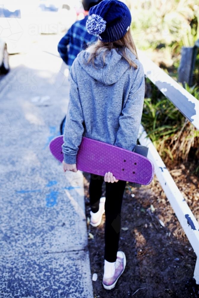 Girl walking away holding skateboard behind her back - Australian Stock Image