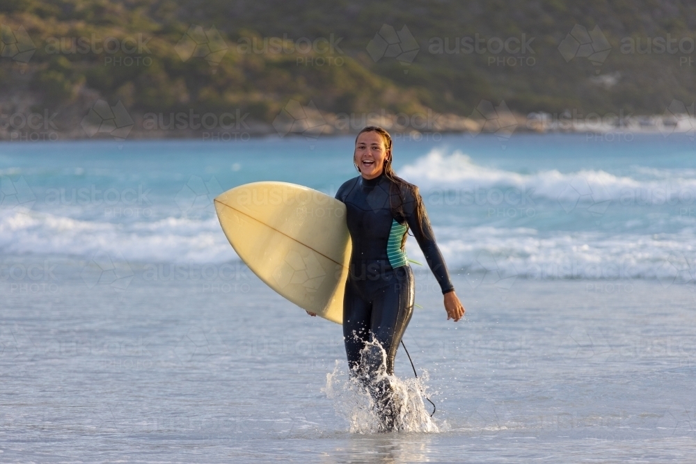 girl walking out of the water carrying surfboard on beach - Australian Stock Image