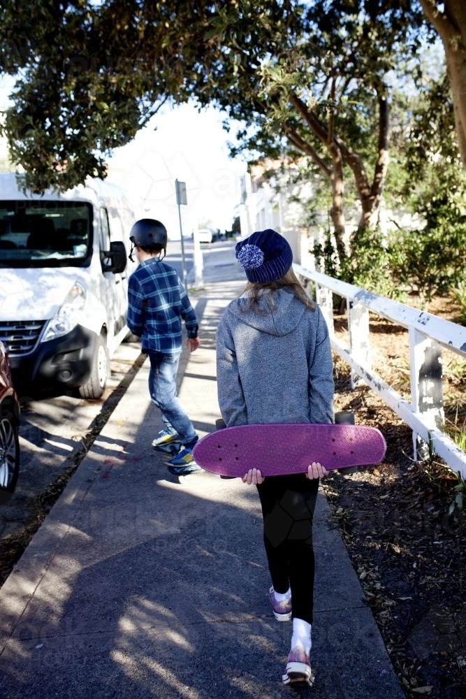 Girl walking up path holding skateboard with boy skating in front of her - Australian Stock Image