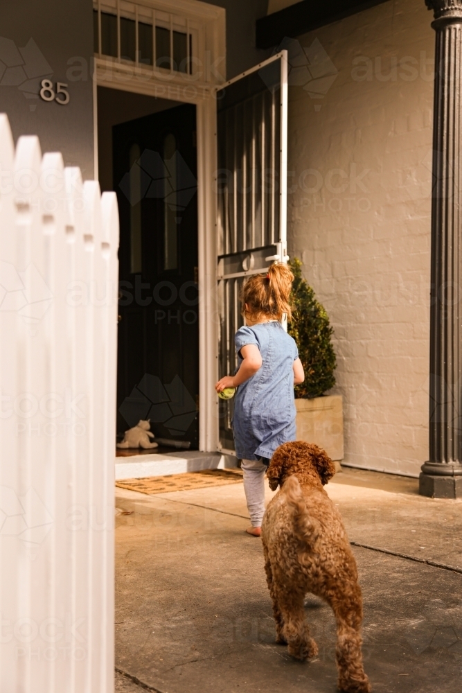 girl wearing blue dress with brown poodle dog running towards the front door of home - Australian Stock Image