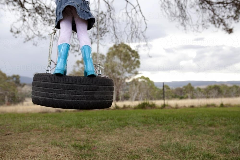 Girl wearing blue gumboot swinging on tyre swing in a country backyard - Australian Stock Image