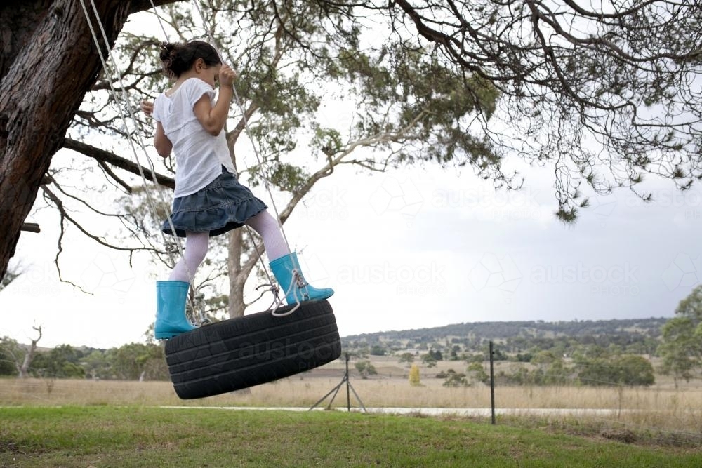 Girl wearing blue gumboot swinging on tyre swing in country backyard - Australian Stock Image