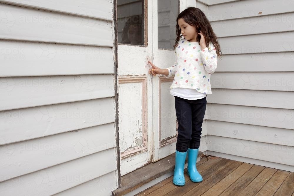 Girl wearing blue gumboots standing by closed door - Australian Stock Image