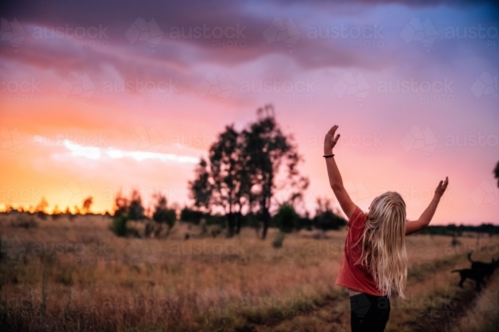 girl with long blonde hair dancing with her hands up in the air with a colourful sky - Australian Stock Image