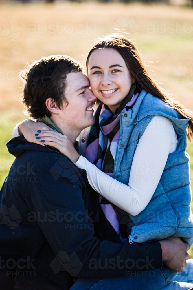 Girlfriend sitting on boyfriends lap with arms around neck smiling - Australian Stock Image