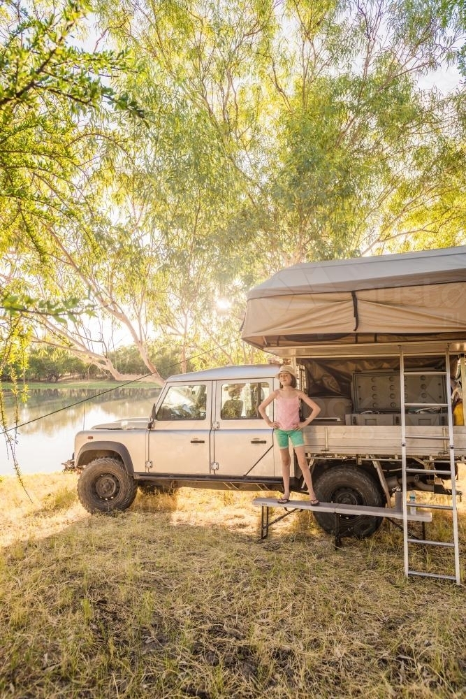 Girls beside four wheel drive with roof top tent by a river in afternoon light - Australian Stock Image