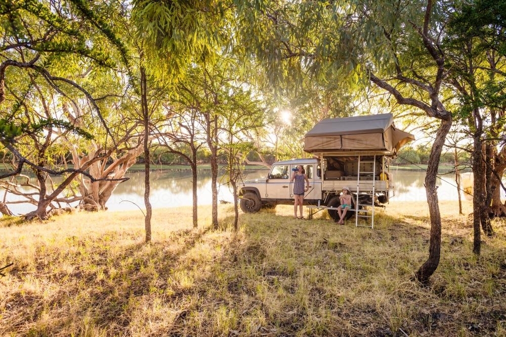 Girls beside four wheel drive with roof top tent by a river in afternoon light - Australian Stock Image