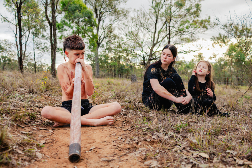 Girls listening to aboriginal boy playing didgeridoo music about dreamtime stories - Australian Stock Image