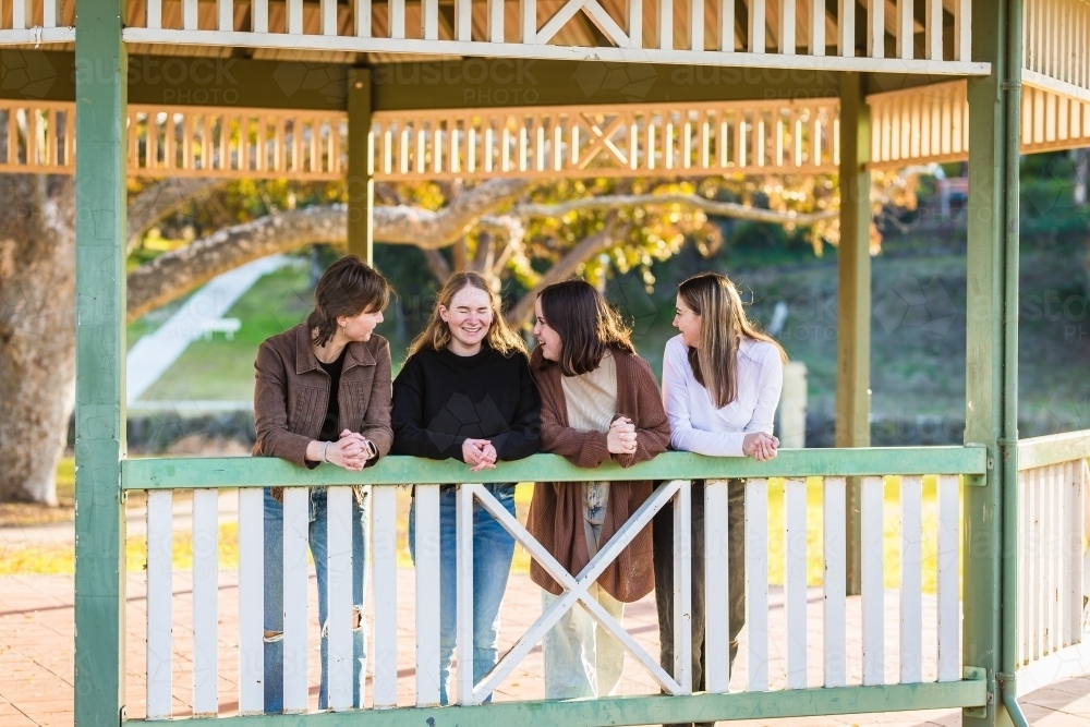 Girls looking at friend laughing leaning on railing of gazebo in park - Australian Stock Image