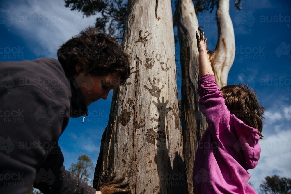 Girls making muddy hand prints on tree, with shadow - Australian Stock Image