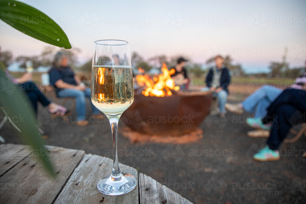 Glass of sparkling wine on a wooden table, with a crowd of women around a firepit in the background - Australian Stock Image