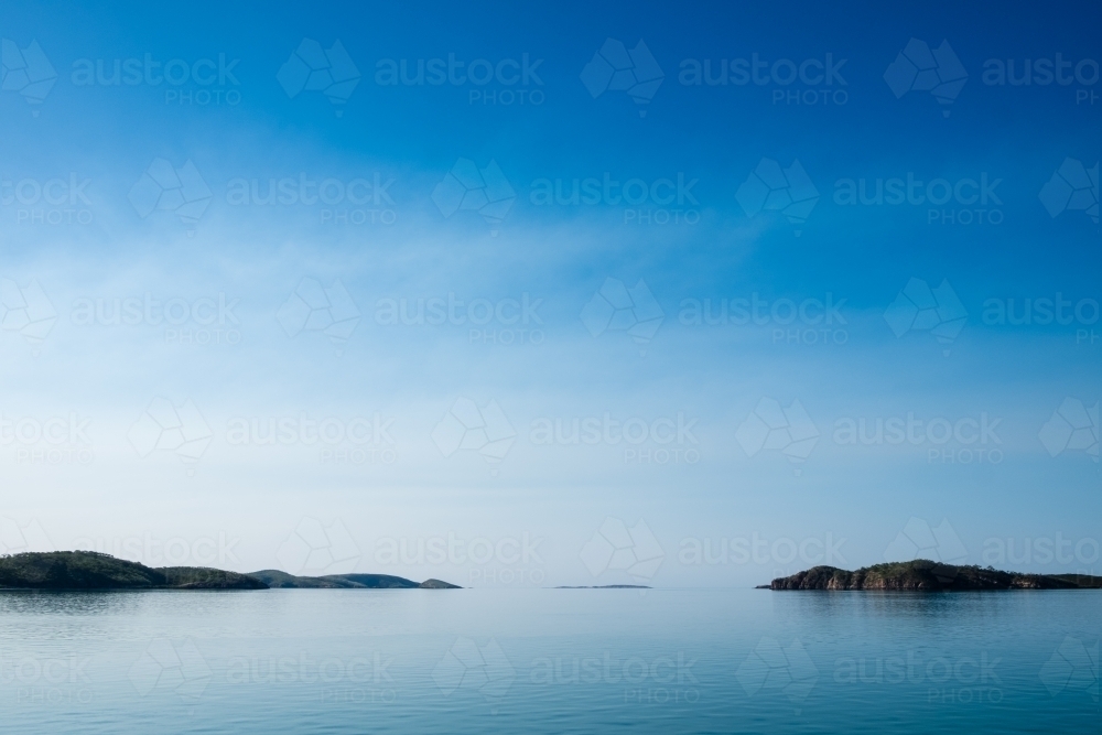 Glass off ocean with islands in distance, horizon blending into sky - Australian Stock Image