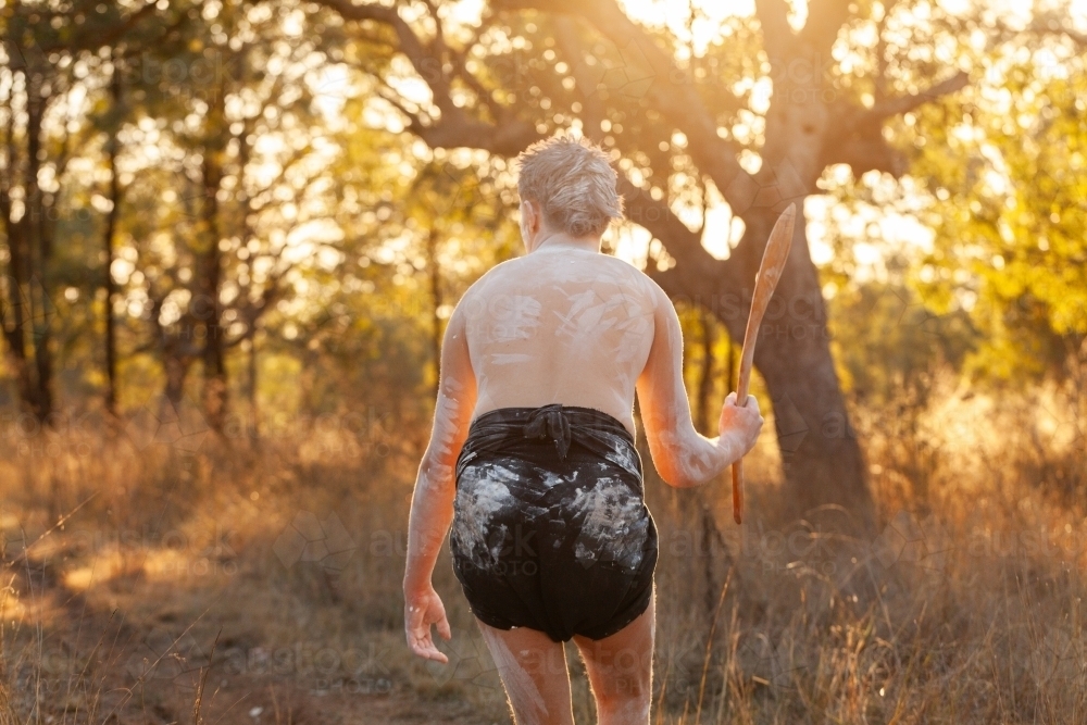 Golden Australian sun behind young First Nations man holding hunting stick in bushland - Australian Stock Image