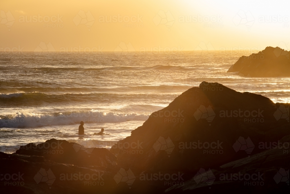 Golden colours of sunlight around the headland of Cabarita with two people in the surf. - Australian Stock Image