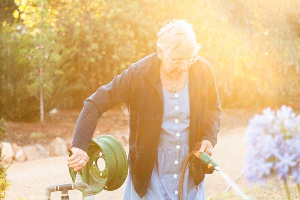 Golden light flare over old woman turning off tap for watering garden - Australian Stock Image