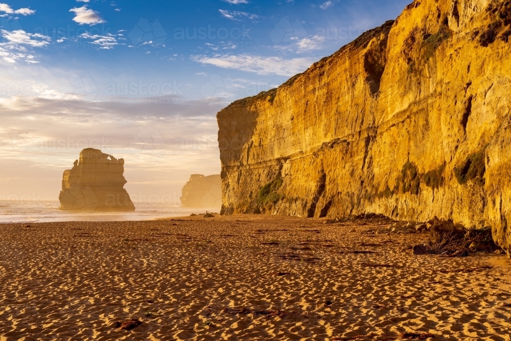 Golden light on the face of a vertical cliff face rising from a sandy beach - Australian Stock Image