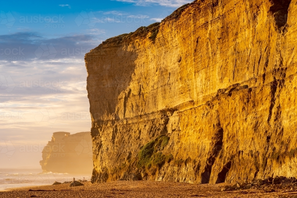 Golden light on the face of a vertical cliff face rising from a sandy beach - Australian Stock Image