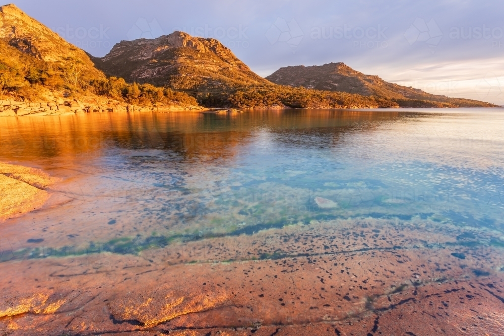 Golden lighting on coastal mountains behind calm water in large rock pools - Australian Stock Image