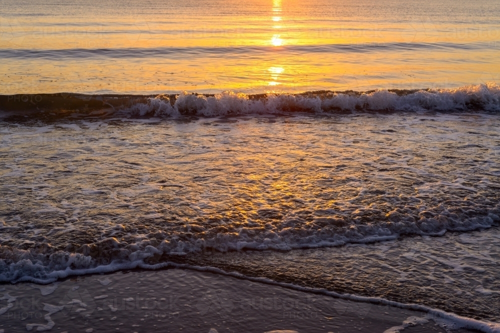 Golden reflections at water's edge at dawn with small waves - Australian Stock Image