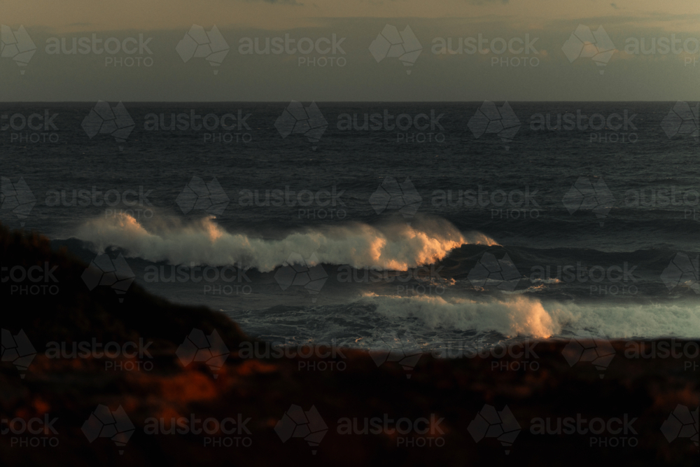 Golden sunlight illuminating ocean waves near rocky shore - Australian Stock Image