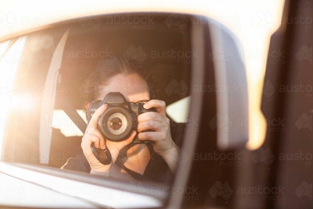 Golden sunlit reflection of photographer on road trip in car passenger mirror - Australian Stock Image