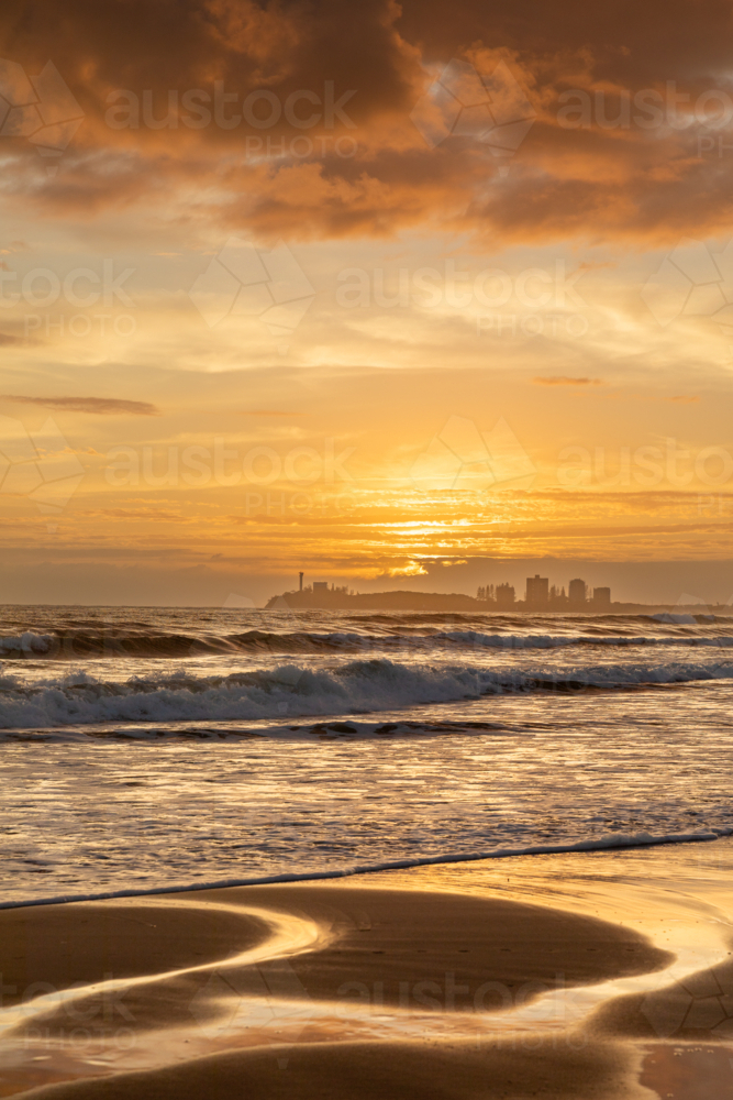 Golden sunrise at the beach with sand reflections, waves and buildings in background - Australian Stock Image