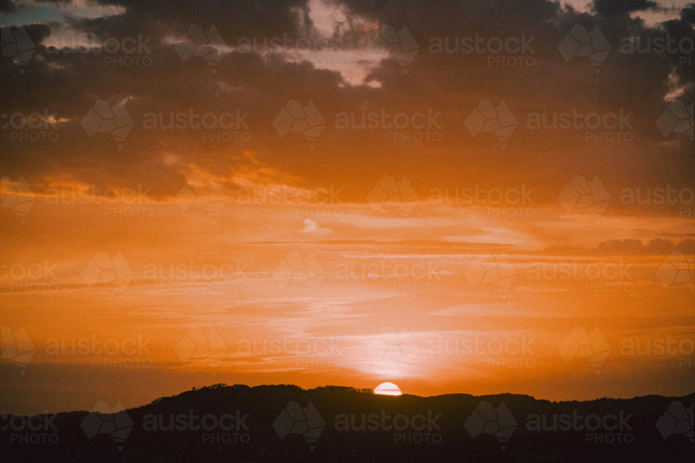 Golden sunset dipping below distant hills beneath dramatic clouds - Australian Stock Image