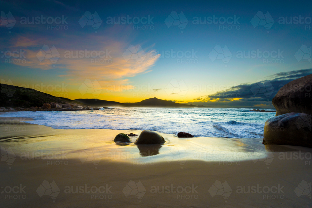 Golden sunset light reflecting off wet sand at a secluded beach with gentle waves and rocks - Australian Stock Image