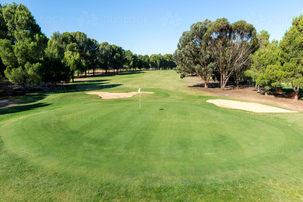 Golf course green and bunkers on a sunny day, surrounded by trees and blue sky - Australian Stock Image
