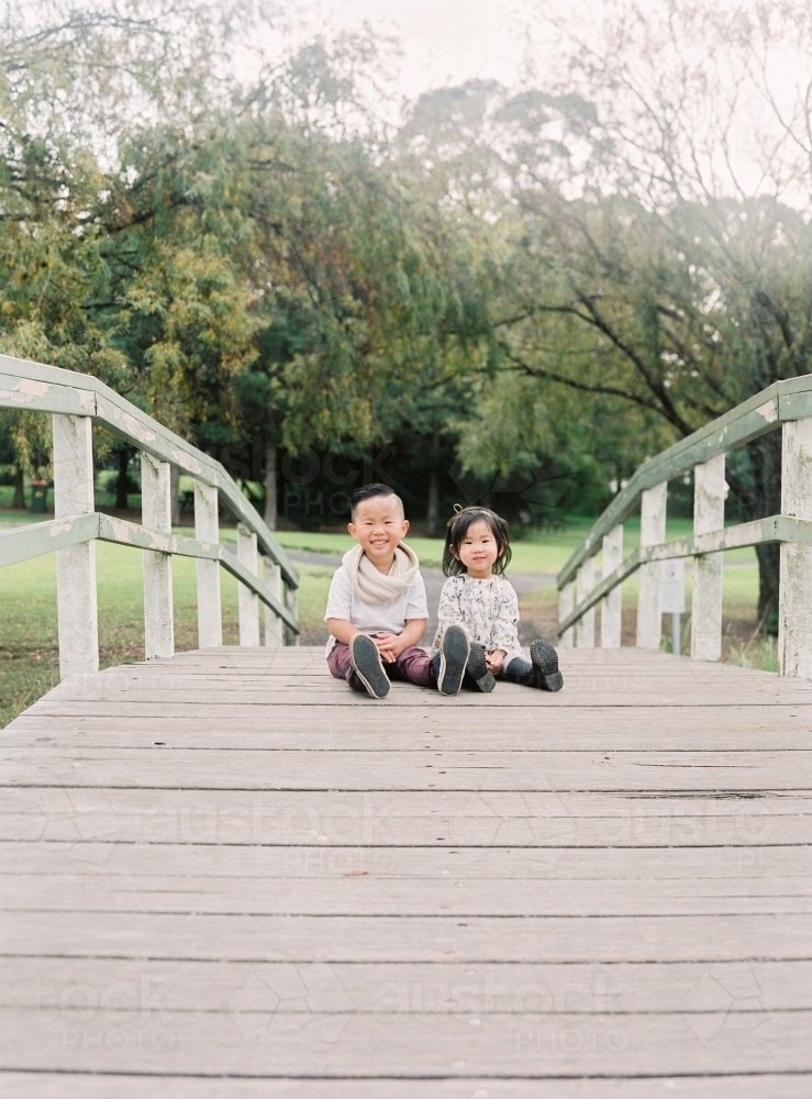 Gorgeous young sister and brother sitting on a footbridge, smiling at the camera - Australian Stock Image
