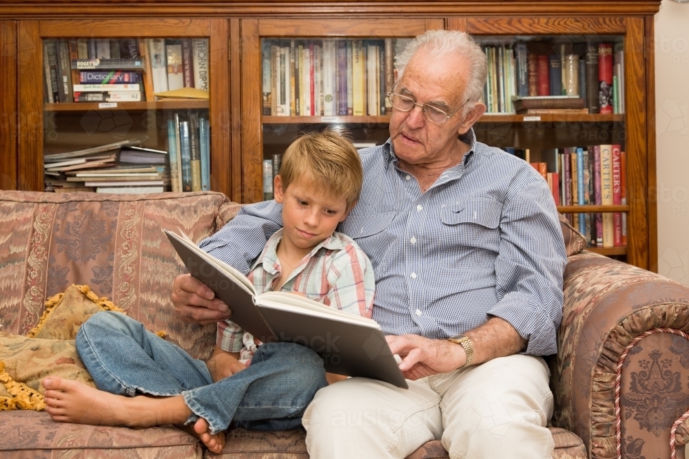 Grandfather and grandson reading a book on a sofa together - Australian Stock Image