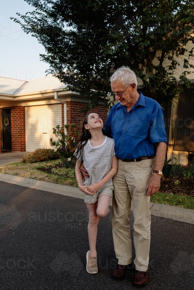 Grandfather hugs his granddaughter as she looks up at him adoringly in front of his house, Australia - Australian Stock Image