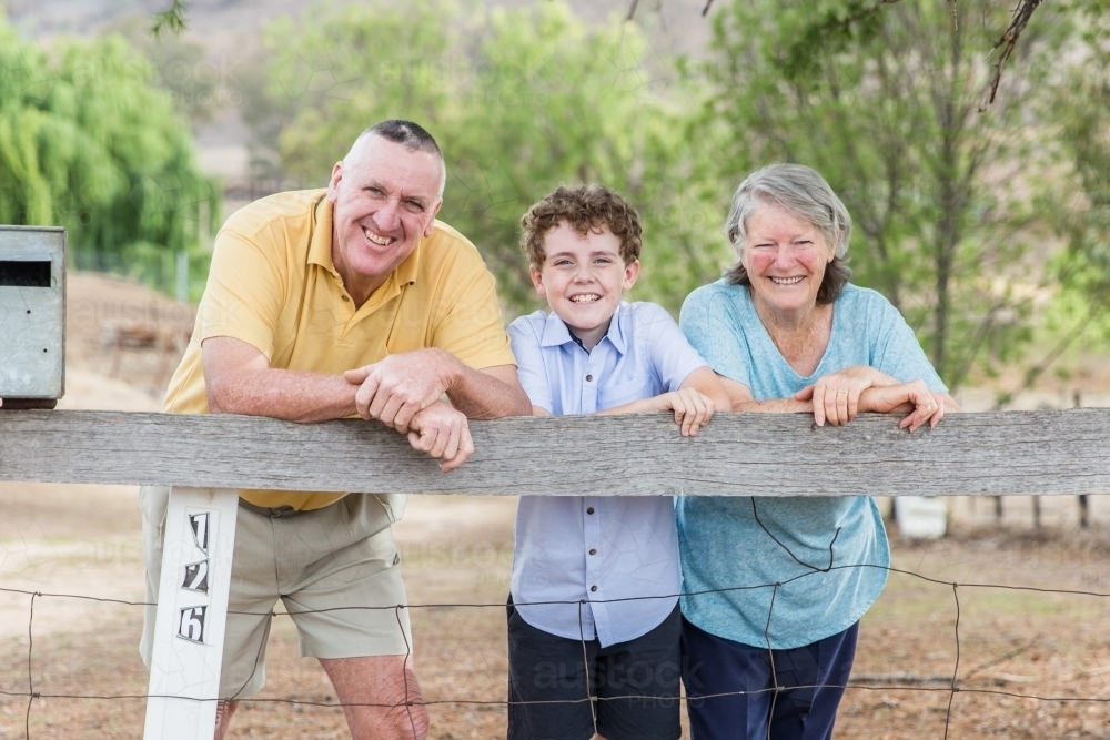 Grandson standing with retired grandparents leaning on front fence at home on farm smiling - Australian Stock Image