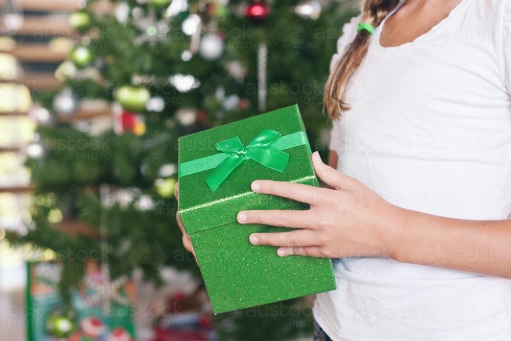 green christmas box in young girl's hands, with blurred xmas tree in the background - Australian Stock Image