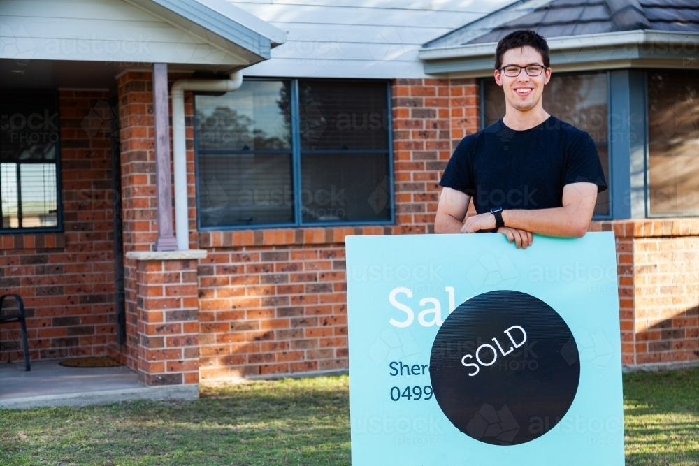 grinning young man with sold sign in front of newly purchased house - Australian Stock Image