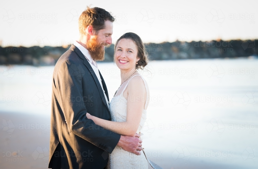 Groom looks at bride as bride looks at the camera outdoors at a beach wedding. - Australian Stock Image