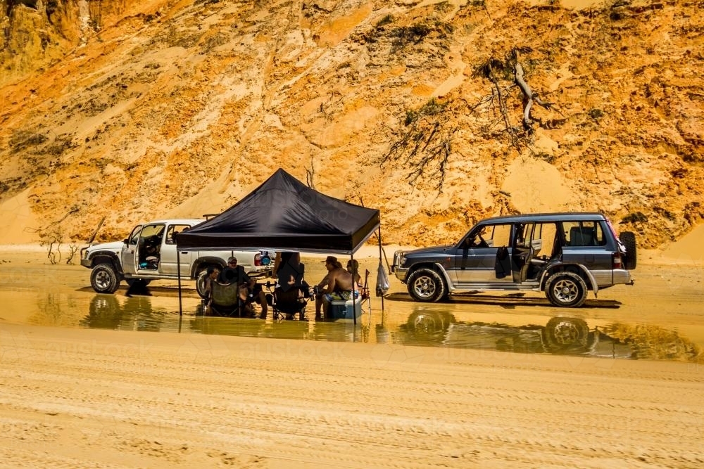 Group of people sit in shallow water beside four wheel drive vehicles - Australian Stock Image