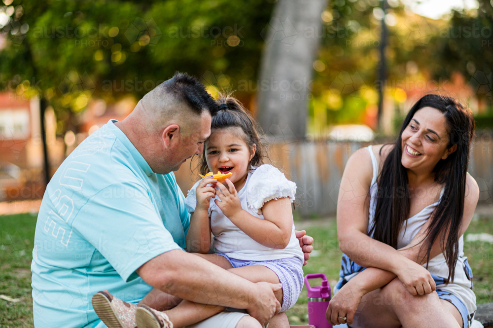 Group of three people share food and laughter while sitting on grass in a park - Australian Stock Image