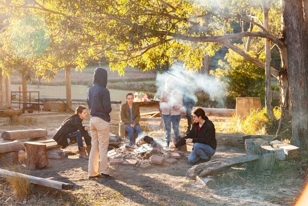 Group of young people relaxing around campfire in bushland on sunny winter morning - Australian Stock Image