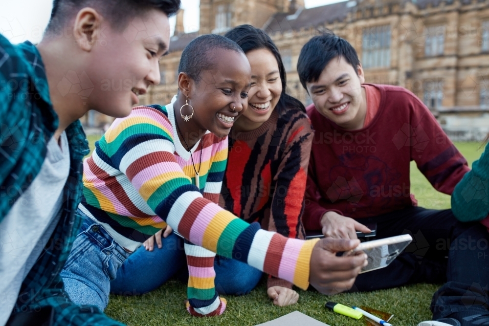 Group of young university students hanging out on grass studying and viewing mobile device - Australian Stock Image